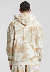 Beige og krem camouflage hettegenser med en avslappet passform, teksturert stoff og ribbestrikkede ermer. Har en kengurulomme.