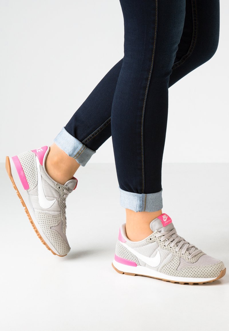 Henkilö, joka on pukeutunut vaaleanharmaisiin ja vaaleanpunaisiin Nike-tennareihin, joissa on valkoinen swoosh-logo, ja hänellä on tummansiniset, kynnykselle käärityt farkut. Hän seisoo valkoisella lattialla.