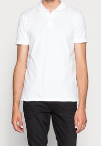 Polo blanc en coton, avec un col classique, des manches courtes et un logo discret sur la poitrine. Associé à un jean foncé.