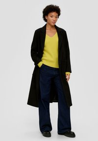 Zwarte lange jas over een gele geribbelde trui en een donkerblauwe wijde jeans. Zwarte plateauschoenen maken de outfit compleet.