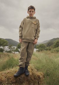 Khaki bluza z kapturem z siateczkową kieszenią, zestawiona z dopasowanymi spodniami dresowymi. Czarne botki za kostkę z sznurowadłami i teksturowaną powierzchnią.