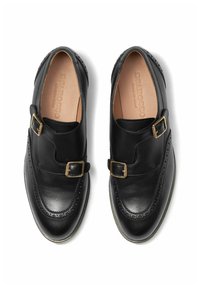 Scarpe in pelle nera con un design a doppia fibbia, dettagli brogue lungo i bordi e una texture liscia con un rivestimento di colore chiaro.