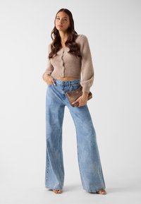 Kort beige vest van een wolmix met knoopdetails, gecombineerd met lichtblauwe jeans met wijde pijpen. Accessoire: bruine clutch met textuur.