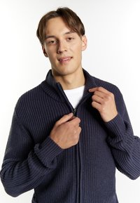 Maglione zip-up in maglia blu navy con texture a coste, colletto alto e chiusura con zip; il modello afferra la zip con entrambe le mani.