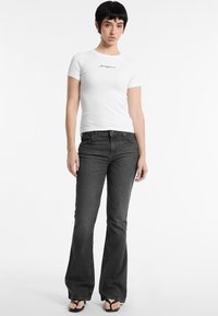 T-shirt blanc à manches courtes avec texte noir, associé à un jean évasé gris foncé. Le modèle portant des sandales noires se tient contre un fond uni.