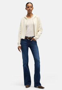 Ljusbeige zip-up hoodie över en figurnära krämfärgad topp, kombinerad med mörkblå utsvängda jeans och brunt bälte. Modellen står mot en neutral bakgrund.