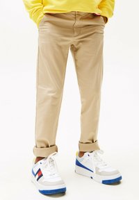Chinos en coton beige avec une texture lisse, aux poignets retroussés, associés à des baskets blanches avec des accents bleu et rouge et une semelle texturée bleue.