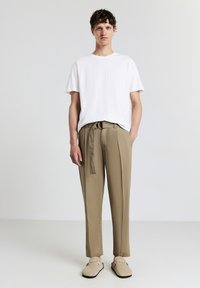 Maglione bianco a maniche corte con motivo testurizzato, abbinato a pantaloni beige con vita legata. Il modello è posizionato su uno sfondo neutro.