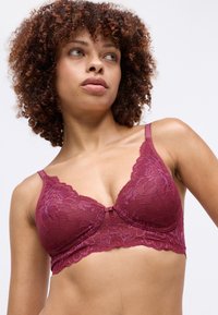 Donna con capelli ricci che indossa un reggiseno in pizzo bordeaux, guardando leggermente di lato su uno sfondo chiaro e semplice.