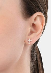 Los pendientes de cruz plateados presentan acentos de perlas redondas, con un tamaño aproximado de 1.5 cm. Se muestran en la oreja de un modelo contra un fondo neutro.