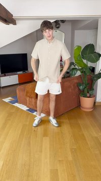 Camisa corta de manga corta color beige claro, pantalones cortos blancos, zapatillas de deporte plateadas. De pie en una habitación con suelo de madera y una planta en maceta.