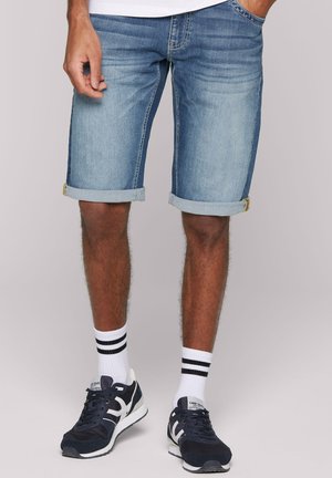 Man wearing light blue rolled-up denim shorts, white socks with black stripes, and navy blue sneakers standing against a plain background.