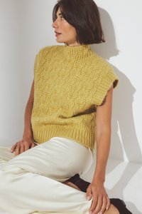 Geel gebreide mouwloze trui met een hoge hals, gekenmerkt door een textuurpatroon. Draagt over witte satijnen broek.
