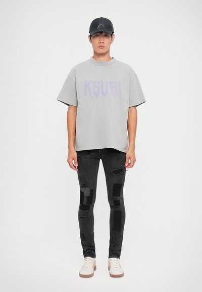 Grå, oversized t-skjorte med lila "KSUBI" grafikk, sammen med utslitte svarte jeans med lapper, og kremfargede sneakers.