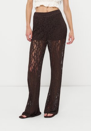 Kvinder iført sorte gennemsigtige blondebukser med vide ben og indbyggede shorts samt en hvid cropped top, stående mod en ensfarvet baggrund.