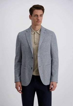 Man staand, gekleed in een blauw gebloesd colbert over een beige poloshirt en donkere broek, tegen een effen lichte achtergrond.