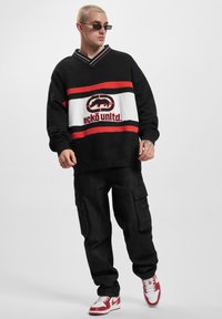 Black oversized sweatshirt with red and white horizontal stripes, featuring a brand logo. Paired with black cargo pants and red sneakers.