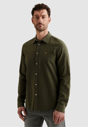 Mand iført en tætsiddende olivengrøn skjorte med lange ærmer og knapper, med et lille broderet emblem på brystet, stående foran en ensfarvet baggrund.