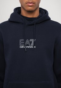 Tamsiai mėlynas medvilnės mišinio džemperis su kapišonu, turintis kengūrinę kišenę ir raištelius. Priekyje yra baltas EA7 Emporio Armani logotipas.
