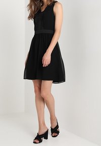 Černé bezramínkové šaty s wovaným detailem na korzetu, rozšířenou sukní a zúženým pasem. Vytvořené v kombinaci s černými sandály na podpatku se uzlovými akcenty.