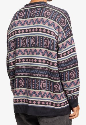 Mężczyzna noszący wielokolorowy sweter dziergany z geometrycznymi i spiralnymi wzorami w odcieniach różu, niebieskiego, czerni i kremu, widziany od tyłu.