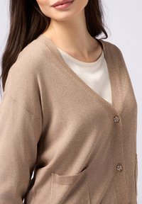 Beige stickad cardigan med V-hals, framfickor och dekorativa blomknappar. Materialet har en subtil lyster.
