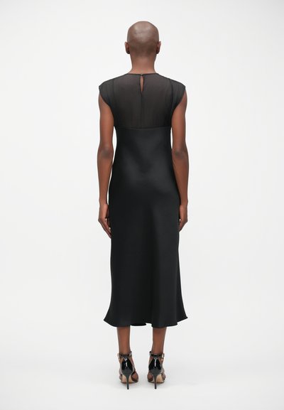 Μαύρο σατέν φόρεμα με διαφανή πλέγμα στην πλάτη, λεπτομέρεια σε σχήμα κλειδώματος και ροή σιλουέτας με ελαφρώς φουσκωτό τελείωμα.