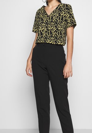 Frau trägt schwarze, maßgeschneiderte Hose und eine schwarze Kurzarmbluse mit gelbem Blumenmuster, steht vor einem schlichten hellen Hintergrund.