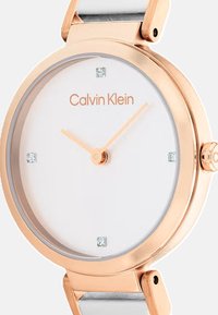 Montre en or rose avec un cadran blanc, design minimaliste, deux aiguilles dorées, quatre petits cristaux et un bracelet en métal avec un accent blanc.