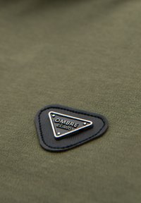 Olivgrön tyg med en smidig textur, med en triangulär svart läderlapp med silverbokstäver "OMBRE EXIMIO."