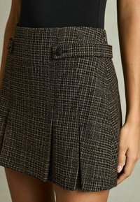 Fekete és barna houndstooth mintás szoknya pliszével, két elülső gombbal, derékrésszel és texturált anyaggal. Közeli felvétel az anyag részleteiről és mintázatáról.