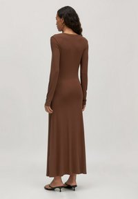 Frau steht mit dem Rücken zur Kamera und trägt ein enges, langärmeliges, braunes Maxikleid sowie schwarze, flache Sandalen vor einem einfarbigen Hintergrund.