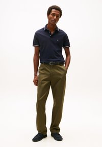 Marinblå pikétröja med vit och röd kragkant, kombinerad med olivgröna byxor och mörkblå skor. Enkel design med avslappnad passform.