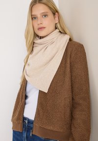 Une écharpe beige drapée autour du cou, assortie à une veste marron texturée. Le tissu semble doux, avec un design décontracté et superposé.