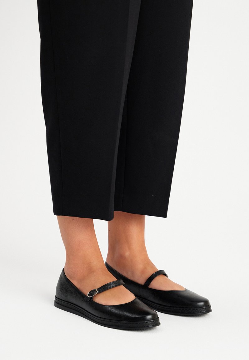Zapatos planos de charol negros estilo Mary Jane con punta redondeada y una correa sobre el empeine, colocados sobre un fondo claro, combinados con pantalones negros cortos.