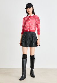 Crvena cardigan jakna s bijelim tekstom, bijelim ovratnikom; crna plisirana suknja s bijelim prugama; crne čizme do koljena s potpeticom, crna kapa.