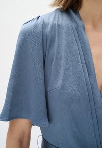 Hellblaues Kleid mit strukturiertem Stoff, kurzen flatternden Ärmeln, einem Wickeldesign und einem dezenten Taschendetail im Brustbereich.
