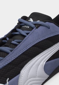 Κοντινό πλάνο μαύρου και μωβ αθλητικού παπουτσιού Puma με λευκό λογότυπο, που αναδεικνύει υφασμάτινη υφή και λεπτομερή ραφές κατά μήκος της πλευράς και των κορδονιών.