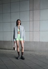 Grå rutig skjorta, ljusgröna träningsshorts, pastellrosa handväska och bruna strumpor med mörka sneakers. Står mot en kaklad vägg.