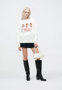 Witte sweater met peperkoekkoekjesgrafieken en rode tekst, gecombineerd met een zwarte mini-rok, zwarte kniehoge laarzen en een kleine witte handtas.