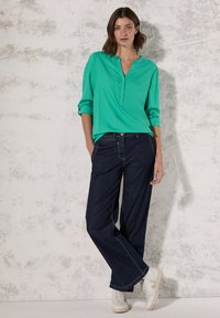 Blusa azzurra con bottoni, a maniche tre quarti, abbinata a jeans a gamba larga blu scuro e sneakers bianche. Sfondo strutturato.