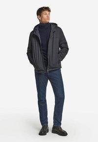 Ung man iklädd en mörkgrå huva-jacka, marinblå polotröja, blå jeans och svarta snörskor, stående med händerna i fickorna.