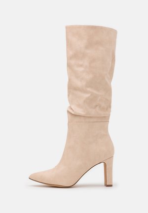 Beige suède kniehoog laars met een slungelige schacht, puntige neus en blokhakken tegen een witte achtergrond.