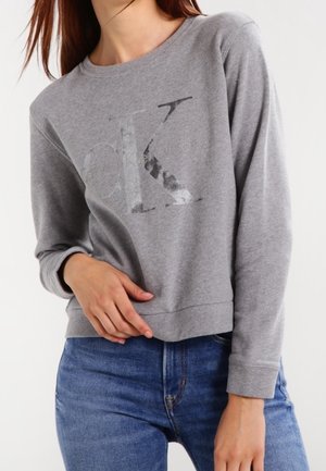 Kobieta w szarej bluzie z długim rękawem z subtelnym logo Calvin Klein i niebieskich dżinsach, z jedną ręką opartą na talii.