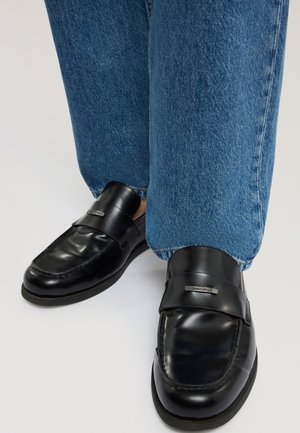 Coppia di mocassini neri in pelle con dettaglio metallico indossati con jeans blu a gamba dritta, mostrati dalla metà del polpaccio in giù su sfondo neutro.