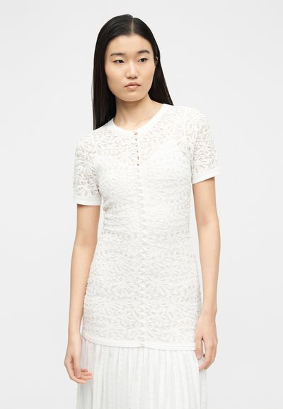 Νεαρή γυναίκα με μακριά σκούρα μαλλιά, φορώντας λευκό δαντελένιο τοπ με κοντά μανίκια και κουμπιά από μαργαριτάρια, και λευκή πλισέ φούστα, στραμμένη ελαφρώς αριστερά.