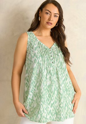 Frau trägt eine ärmellose grün-weiß gemusterte Bluse mit Knöpfen und Falten, steht vor einem schlichten beigen Hintergrund.