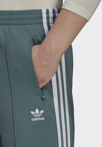 Pantaloni da pista Adidas in tessuto verde con strisce laterali bianche, tasca con zip e logo ricamato sul davanti. Texture liscia.