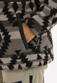 Veste en polaire avec un motif géométrique gris et noir, dotée d'une poche sur le côté, d'une texture douce et d'une bordure noire contrastante.