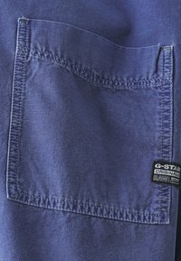 Blått tyg med en texturerad yta, med en rektangulär ficka och en G-Star Originals svart etikettsöm på höger sida.